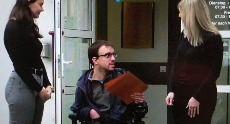 Zwei Auszubildende der Kreisverwaltung stehen am Hinterausgang der Kreisverwaltung. In ihrer Mitte ist ein Rollstuhlfahrer zu sehen, der ein Formular übergibt.