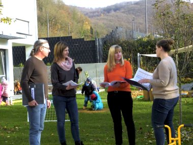 Eine Mitarbeiterin und ein Mitarbeiter der Kreisverwaltung und zwei Mitarbeiterinnen einer KiTa stehen im Garten der KiTa und besprechen etwas. Im Hintergrund sieht man spielende Kinder.