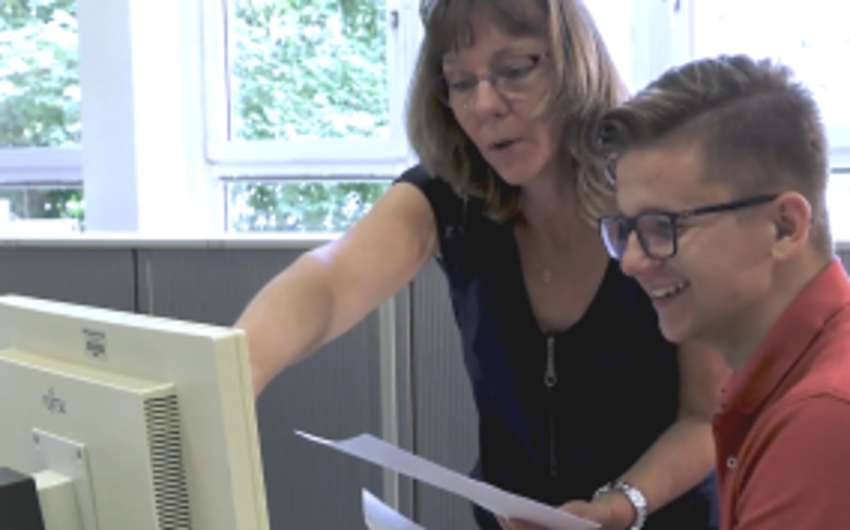 Eine Mitarbeiterin erklärt einem Auszubildenden etwas am Computer.