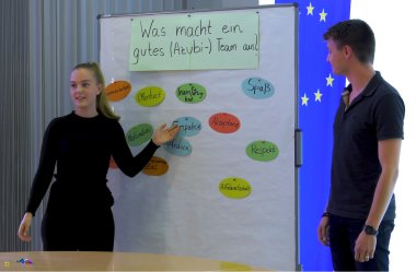 Eine Auszubildende steht an einer Präsentationswand auf der mehrere Zettel angebracht sind. Rechts daneben steht ein Auszubildender.