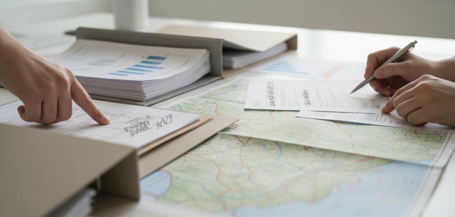 Sachliche Szene mit Förder- und Projektunterlagen auf einem Tisch. Neutrale Aktenmappen, lose Dokumente und Kartenmaterial einer Region liegen ausgebreitet nebeneinander. Ein Zuwendungsbescheid bzw. Förderunterlagen sind erkennbar, jedoch ohne lesbare Schrift oder Logos. Dezenter Fokus auf Zahlen, Planung und Zusammenarbeit. Helle, ruhige Lichtstimmung, moderner Verwaltungs- und Planungskontext. Dokumentarischer Stil, natürliche Farben, keine Inszenierung, keine Personen oder nur Hände im Bild