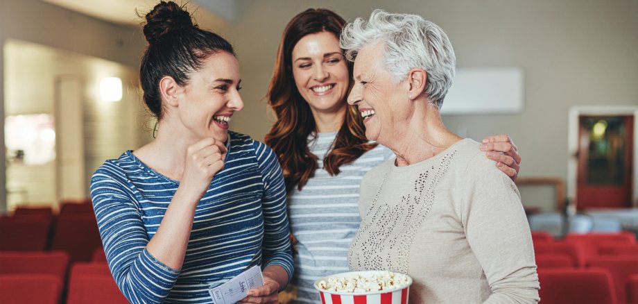 Realistische, dokumentarische Fotografie von drei Frauen aus unterschiedlichen Generationen (eine junge Frau Anfang 20, eine Frau mittleren Alters, eine ältere Frau etwa 70 Jahre), die gemeinsam in einem kleinen Programmkino stehen. Sie lachen und unterhalten sich, halten Popcorn und Kinokarten in der Hand. Warmes Vormittagslicht, natürliche Farben, authentische Stimmung, alltägliche Kleidung, ehrliche Emotionen, Momentaufnahme, leichte Tiefenunschärfe, hohe Auflösung, verbindende und solidarische Atmosphäre.