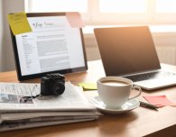 Auf einem Schreibtisch steht ein Computer und ein Laptop inmitten von Pressenotizen und Zeitungen.