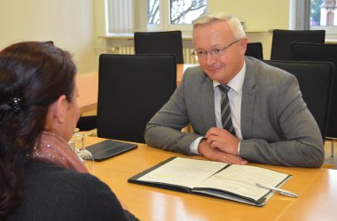 Landrat Achim Hallerbach sitzt hinter einem Schreibtisch und berät eine Bürgerin, die vor dem Schreibtisch sitzt.