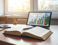 Aufgeschlagenes Buch auf einem Schreibtisch. Dahinter steht ein Laptop.