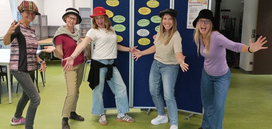 Auf dem Bild sind 5 Frauen zu sehen, die uns die Hände entgegenstrecken und Jede hat einen Hut auf dem Kopf.