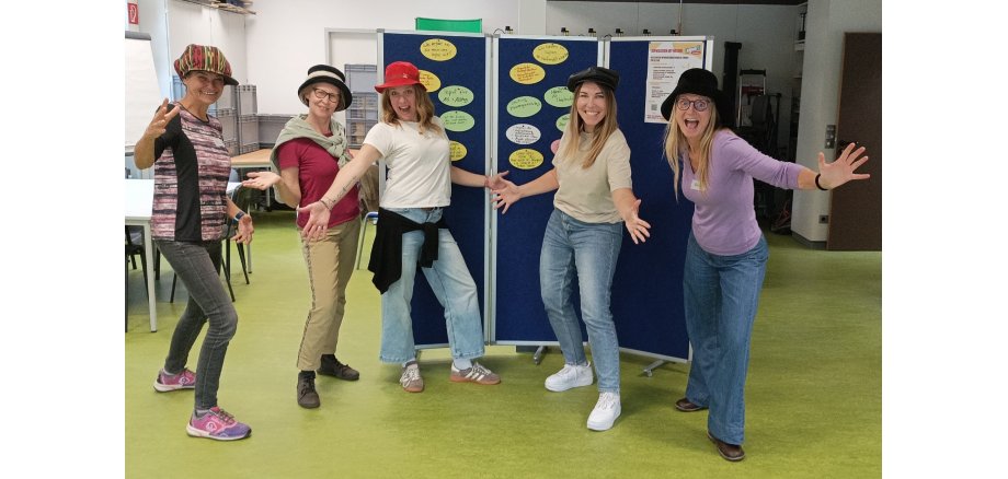 Auf dem Bild sind 5 Frauen zu sehen, die uns die Hände entgegenstrecken und Jede hat einen Hut auf dem Kopf.