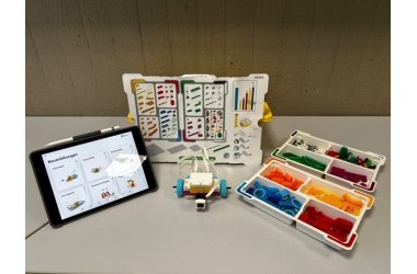 Auf einem Tisch stehen die Utensilien für Lego-Spike: Tablet, Plan mit den einzelen Legobausteinen, Legobausteine und das Anfangsmodell für den Roboter.