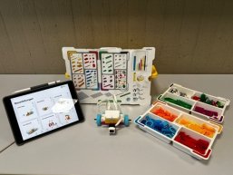 Auf einem Tisch stehen die Utensilien für Lego-Spike: Tablet, Plan mit den einzelen Legobausteinen, Legobausteine und das Anfangsmodell für den Roboter.