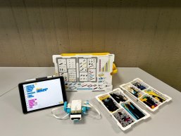 Auf einem Tisch stehen die Utensilien für Lego-Spike: Tablet, Plan mit den einzelen Legobausteinen, Legobausteine und das Anfangsmodell für den Roboter.
